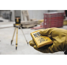 Hand in Arbeitshandschuh hält Fernbedienung für Stabila Rotationslaser auf einer Baustelle mit Stativ im Hintergrund.