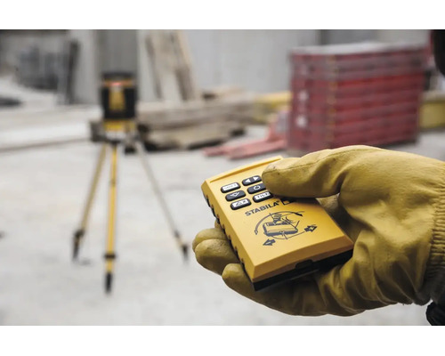Hand in Arbeitshandschuh hält Fernbedienung für Stabila Rotationslaser auf einer Baustelle mit Stativ im Hintergrund.