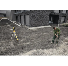 Ein Mann nutzt einen Rotationslaser auf einem Stativ und eine Messlatte mit Empfänger auf einer Baustelle vor einem modernen Gebäude aus Stein.