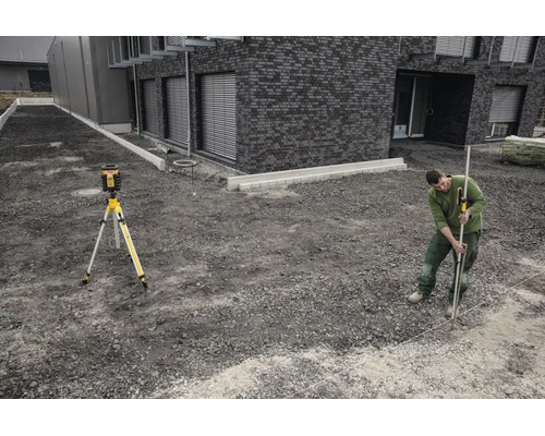 Ein Mann nutzt einen Rotationslaser auf einem Stativ und eine Messlatte mit Empfänger auf einer Baustelle vor einem modernen Gebäude aus Stein.