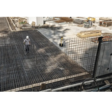 Bauarbeiter auf einer Baustelle mit großflächiger Bewehrung aus Baustahlmatten und einem Vermessungsgerät auf einem Stativ.