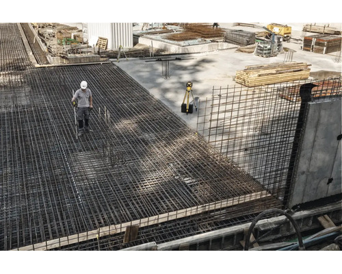 Bauarbeiter auf einer Baustelle mit großflächiger Bewehrung aus Baustahlmatten und einem Vermessungsgerät auf einem Stativ.