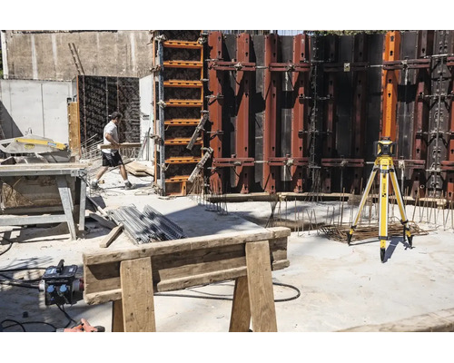 Baustelle mit Stabila Rotationslaser auf Stativ, Schalungswänden aus Metall, Tischkreissäge und einem Bauarbeiter.