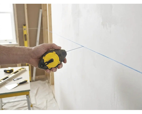 Hand nutzt ein Stanley Schlagschnurgerät für eine blaue Markierungslinie an einer Wand in einer hellen und organisierten Baustellenszenerie.