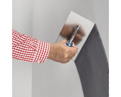 Eine Hand trägt mit einer Zahnkelle Fliesenkleber auf eine Wand auf, wodurch ein gleichmäßiges Rillenmuster entsteht.