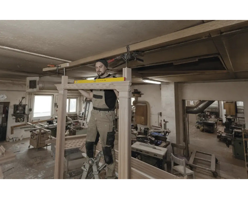 Handwerker in einer Werkstatt prüft mit einer Wasserwaage die Ausrichtung eines Holzrahmens, während er auf einer Leiter steht.