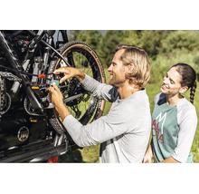 Ein Mann sichert ein Mountainbike mit einem Vorhängeschloss und einem Kabelschloss an einem Fahrradträger am Auto, während eine Frau zusieht.