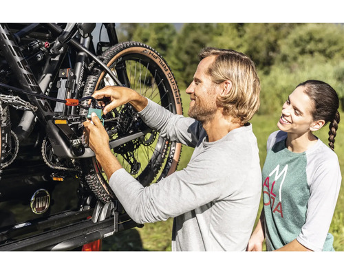 Ein Mann sichert ein Mountainbike mit einem Vorhängeschloss und einem Kabelschloss an einem Fahrradträger am Auto, während eine Frau zusieht.