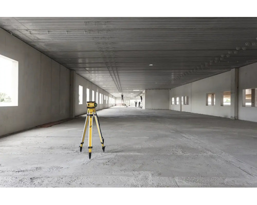 Rotationslaser auf einem Stativ in einem großen, hellen Industrierohbau mit Betonboden und hohen Decken.
