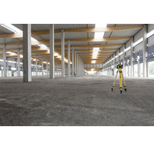 Ein Rotationslaser auf einem Stativ in einer großen, hellen Lagerhalle mit Betonpfeilern und Holzbalken.