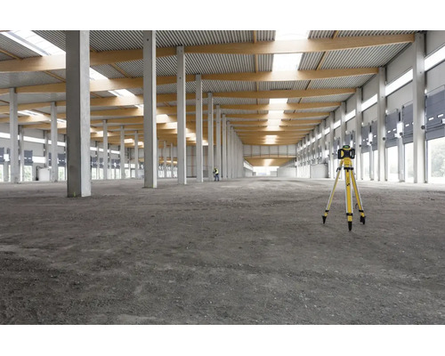 Ein Rotationslaser auf einem Stativ in einer großen, hellen Lagerhalle mit Betonpfeilern und Holzbalken.
