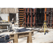 Baustelle mit Stabila Rotationslaser auf Stativ, Schalungswänden aus Metall, Tischkreissäge und einem Bauarbeiter.