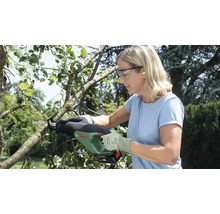 Eine Frau schneidet mit einer Bosch Akku-Gartensäge einen Ast im Garten und trägt dabei Schutzbrille sowie Handschuhe.