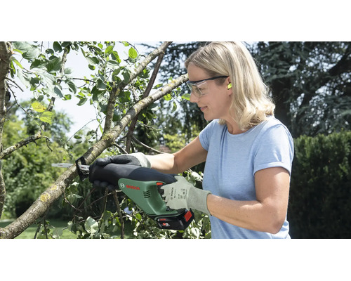 Eine Frau schneidet mit einer Bosch Akku-Gartensäge einen Ast im Garten und trägt dabei Schutzbrille sowie Handschuhe.