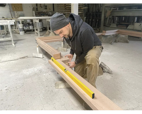 Ein Handwerker kniet in einer Werkstatt und überprüft die Ausrichtung eines großen Holzrahmens mit einer gelben Wasserwaage.