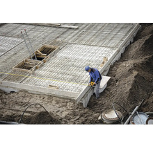 Bauarbeiter auf einer Baustelle misst ein Betonfundament mit Stahlbewehrung mit einem gelben Maßband.