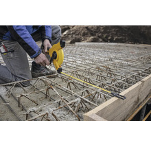 Bauarbeiter auf Bewehrungsstahl misst mit einem gelben Rollmaßband den Abstand zu einer Schalung aus Holz auf einer Baustelle.