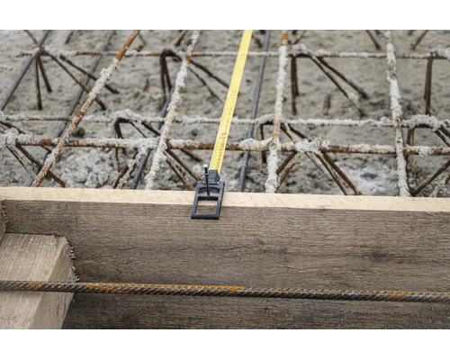 Maßband an Holzschalung fixiert über Bewehrungsstahl und Beton auf einer Baustelle.