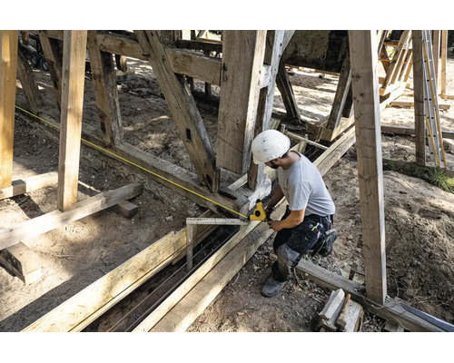 Bauarbeiter mit weißem Schutzhelm misst eine Holzkonstruktion mit einem gelben Maßband auf einer Baustelle.