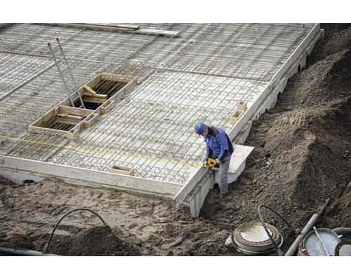 Bauarbeiter auf einer Baustelle misst ein Betonfundament mit Stahlbewehrung mit einem gelben Maßband.