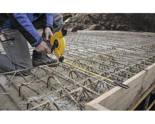 Bauarbeiter auf Bewehrungsstahl misst mit einem gelben Rollmaßband den Abstand zu einer Schalung aus Holz auf einer Baustelle.