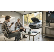Zwei Männer in einer hellen, organisierten Garage beim Betrachten eines Tablets, im Hintergrund ein Auto mit offener Motorhaube und eine Werkzeugwand.