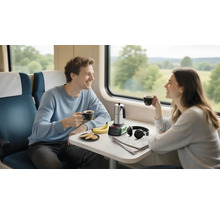 Zwei Personen im hellen Zugabteil an einem Tisch mit mobilem Bosch Espressokocher, Tassen, Keksen, Bananen, Kopfhörern und einer Zeitung.