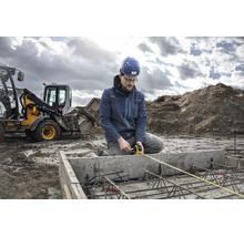 Ein Bauarbeiter mit Schutzhelm misst auf einer Baustelle mit einem Bagger ein Betonfundament mit Bewehrung mittels Maßband.