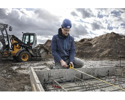 Ein Bauarbeiter mit Schutzhelm misst auf einer Baustelle mit einem Bagger ein Betonfundament mit Bewehrung mittels Maßband.