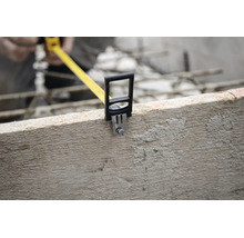 Nahaufnahme eines Maßbandes, das an der Kante eines rauen Holzbretts auf einer Baustelle eingehakt ist.