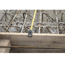 Maßband an Holzschalung fixiert über Bewehrungsstahl und Beton auf einer Baustelle.