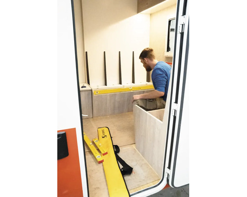 Ein Mann installiert Holzkomponenten mit einer Stabila Wasserwaage in einem hellen Raum mit hellen Holzpaneelen und weißer Wand.