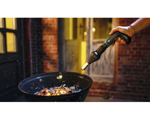 Eine Person verwendet einen Bosch IXO Akkuschrauber mit Grillgebläse-Aufsatz zum Anzünden von Holzkohle in einem Kugelgrill.