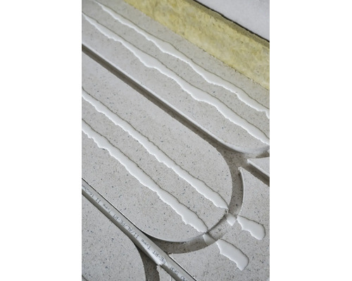 Installation einer Fußbodenheizung mit grauen Systemplatten, weißem Kleber, einem Verbundrohr und Dämmstreifen.