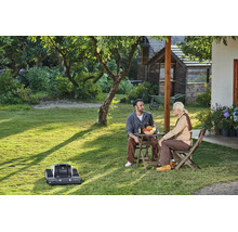 Ein Mähroboter Lymow auf einer Rasenfläche in einem sonnigen Garten, während zwei Männer an einem Tisch sitzen und sich unterhalten.