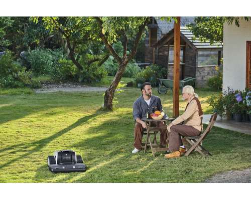 Ein Mähroboter Lymow auf einer Rasenfläche in einem sonnigen Garten, während zwei Männer an einem Tisch sitzen und sich unterhalten.