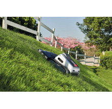 Mähroboter mit Raupenantrieb mäht einen steilen Rasenhang in einem Garten mit weißem Zaun.