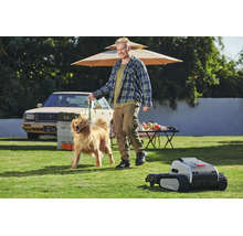 Mähroboter mit Kettenlaufwerk auf einem Rasen mit einem Mann und einem Hund sowie einem Tisch und einem Auto in einem weitläufigen, hellen Garten.