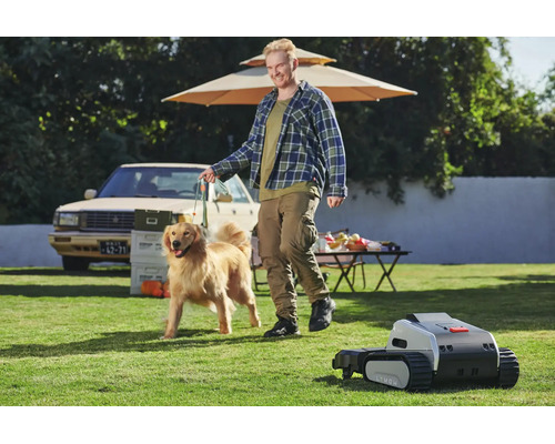 Mähroboter mit Kettenlaufwerk auf einem Rasen mit einem Mann und einem Hund sowie einem Tisch und einem Auto in einem weitläufigen, hellen Garten.