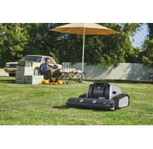 Mähroboter Lymow mit Kettenantrieb auf einer Rasenfläche in einem sonnigen Garten mit Tisch, Sonnenschirm und einer Person im Hintergrund.