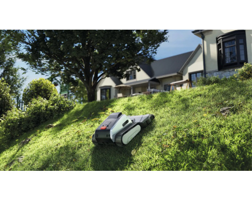Moderner Mähroboter mit Raupenantrieb mäht einen steilen Rasenhang in einem hellen Garten vor einem Wohnhaus.