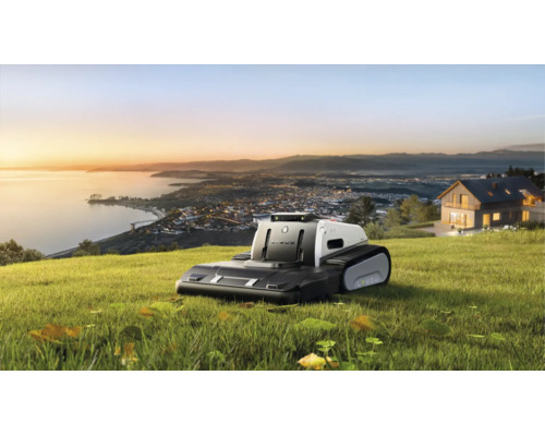 Lymow Mähroboter mit Raupenantrieb auf einer Rasenfläche mit Ausblick auf eine Küstenstadt bei Sonnenuntergang.