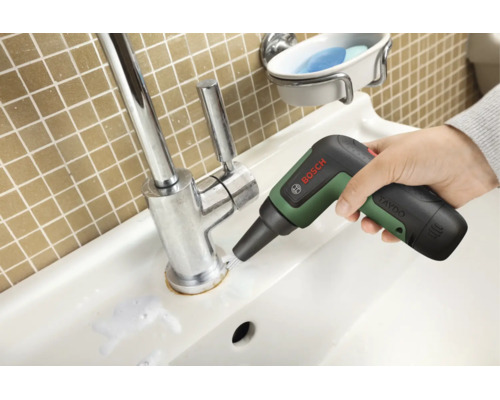 Eine Hand führt die Bosch UniversalBrush Akku-Reinigungsbürste am Sockel einer verchromten Waschbeckenarmatur entlang.