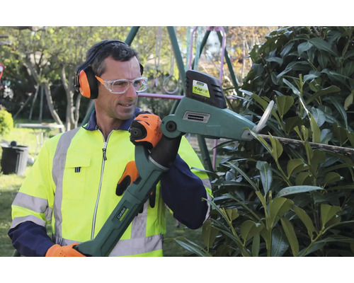 Mann schneidet eine Hecke mit einer motorisierten Heckenschere, trägt Gehörschutz, Schutzbrille, Handschuhe und Warnweste.