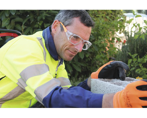 Mann in Warnschutzjacke mit Schutzbrille und Arbeitshandschuhen arbeitet an einem Betonstein im Garten.