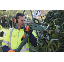 Mann schneidet eine Hecke mit einer motorisierten Heckenschere, trägt Gehörschutz, Schutzbrille, Handschuhe und Warnweste.