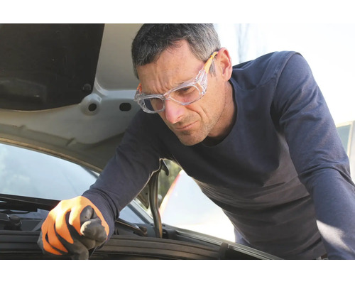 Mann mit Schutzbrille und Arbeitshandschuhen kontrolliert den Motor unter der geöffneten Motorhaube eines Autos.
