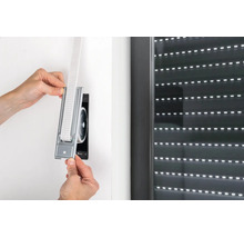 Hände setzen einen Einlass-Gurtwickler mit Gurtband in die Wandöffnung neben einem Rollladenfenster ein.
