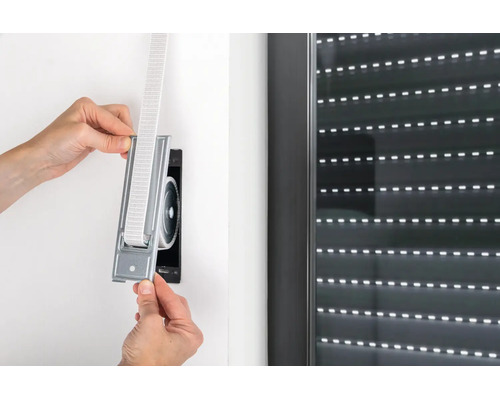 Hände setzen einen Einlass-Gurtwickler mit Gurtband in die Wandöffnung neben einem Rollladenfenster ein.