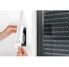 Hände montieren eine weiße Gurtführung für Rolllladengurte an eine Unterputzdose neben einem Fenster mit geschlossenem Rollladen.